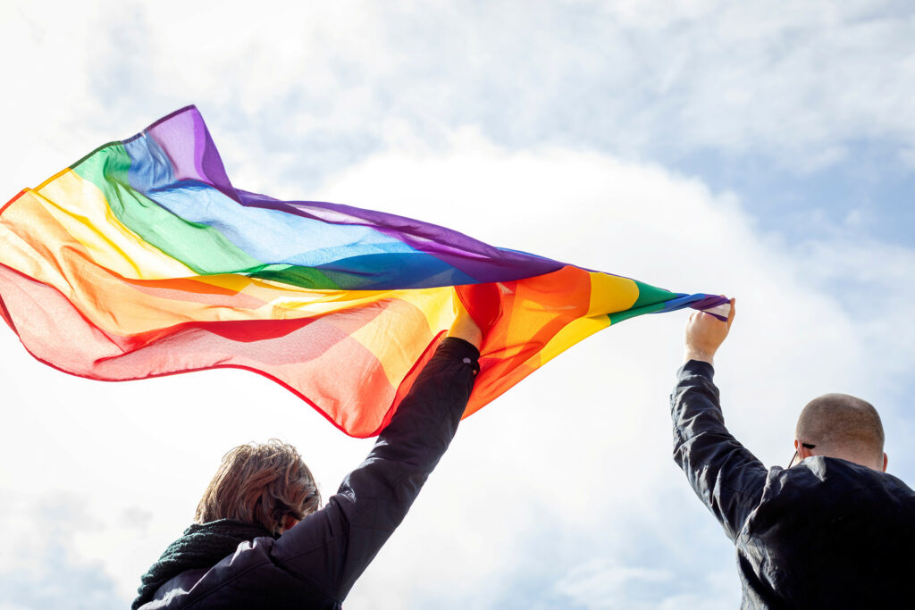Bild på två personers armar som tillsammans håller upp en pride-flagga mot himlen.