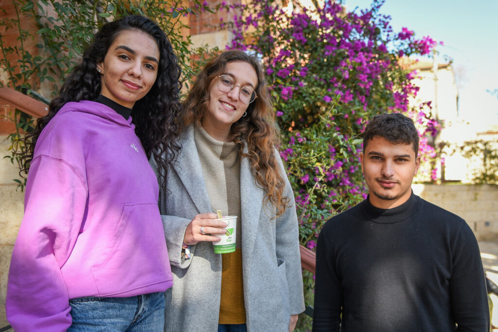 Tre ungdomar i äldre tonåren står bredvid varandra och tittar in i kameran. Längst till vänster står två flickor med långt och lockigt hår, bredvid dem står en pojke med svart tröja.
