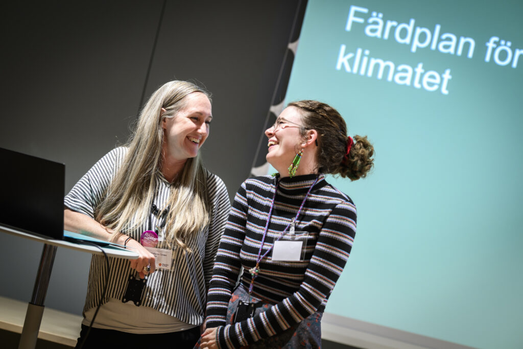 Två kvinnor ler mot varandra. Bakom syns en skärm med texten "Svenska kyrkans färdplan för klimatet".