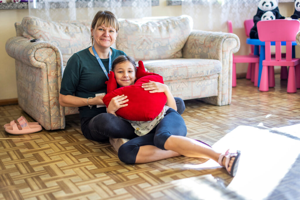 En kvinna och en flicka sitter på golvet, flickan har en röd hjärtkudde i famnen och lutar sig mot kvinnans ben.