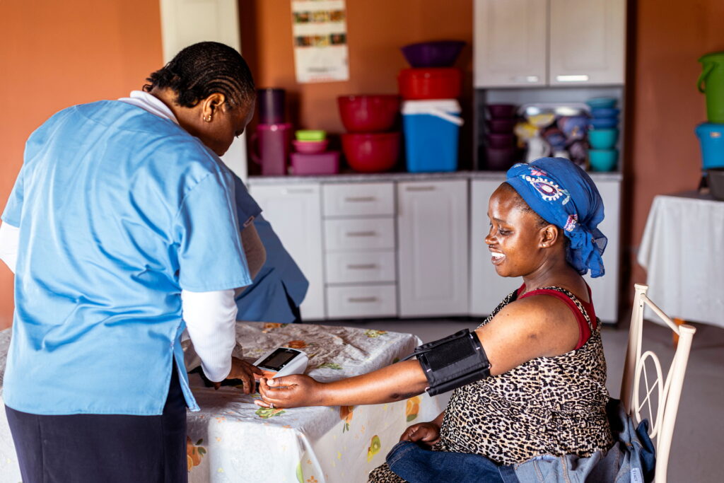 Kvinna står böjd över ett bord. Vid bordet sitter en annan kvinna, hon är glad och har blodtrycksmätare på armen.