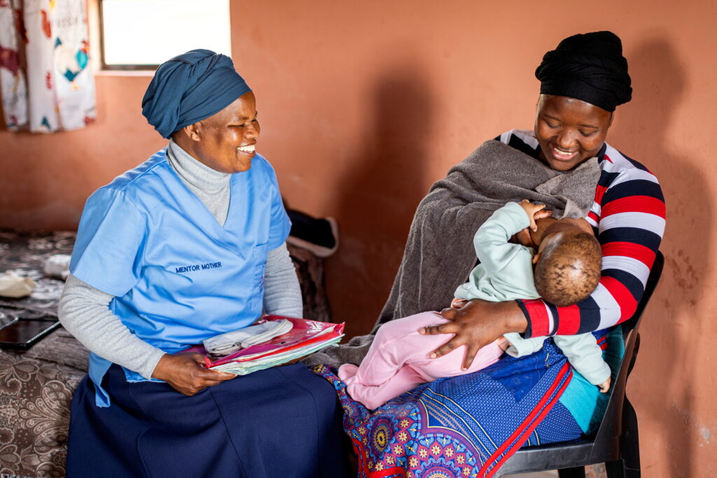 Kvinna sitter och amma bebis. Bredvid sitter kvinna i blå tröja med texten Mentor Mother.