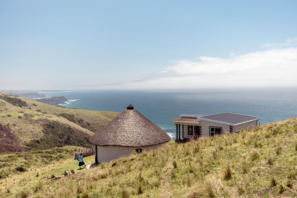 Utsikt över öppet landskap, hav i bakgrunden. I förgrunden två byggnader av enkel standard.