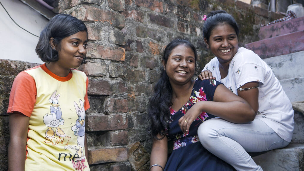 Bild på tre indiska tonårsflickor som sitter tillsammans i en stentrappa. Flickorna tittar på någon utanför bild och skrattar. 
