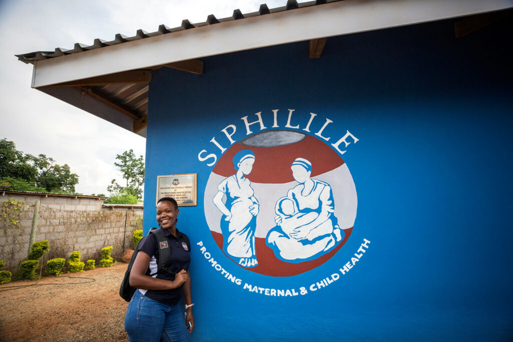 Ung kvinna med ryggsäck och arbetskläder står lutad mot husvägg med stor skylt med logotyp för organisationen Siphilile.