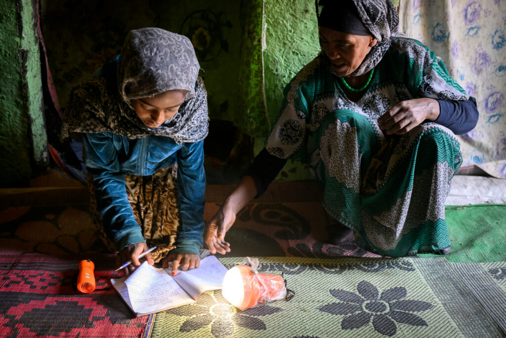 Flicka gör sin läxa i skenet från en solcellslampa. Mamman ger stöd. 