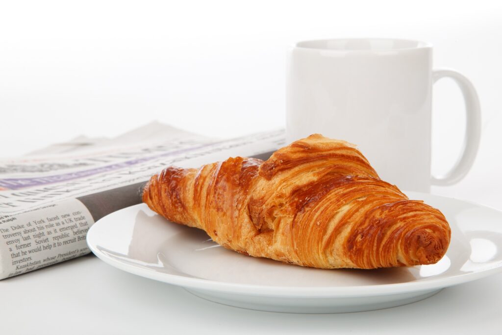 En morgontidning ligger hopvikt intill en kopp kaffe och en croissant.