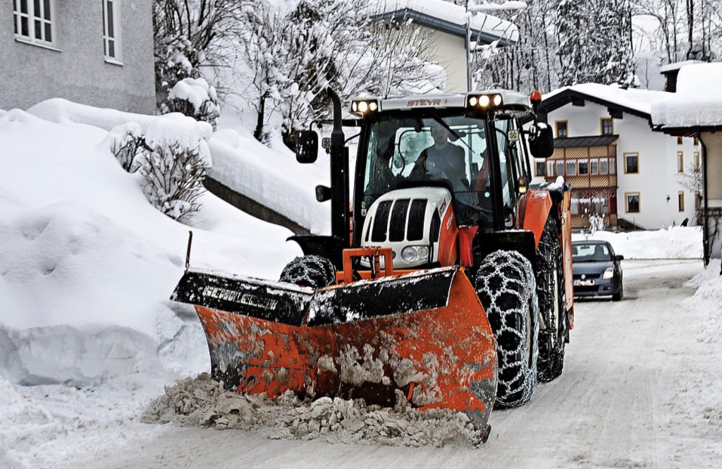 En snöig stadsgata där man ser hus till vänster, och på gatan finns en orange snöplog med kedjor på däcken och ett stort rött plogblad i förgrunden. I bakgrunden en blå personbil. 