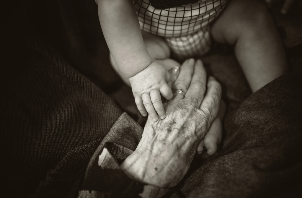 Vi ser en svartvit bild. I övre mitt sträcks en babys hand fram över bebisens ben och den möter en gammal dams hand. Den gamla handen är rynkig och kontrasten mellan händerna tydlig. Den äldre handen har två guldringar på ringfingret. Händerna möts i en stämning av ömhet.