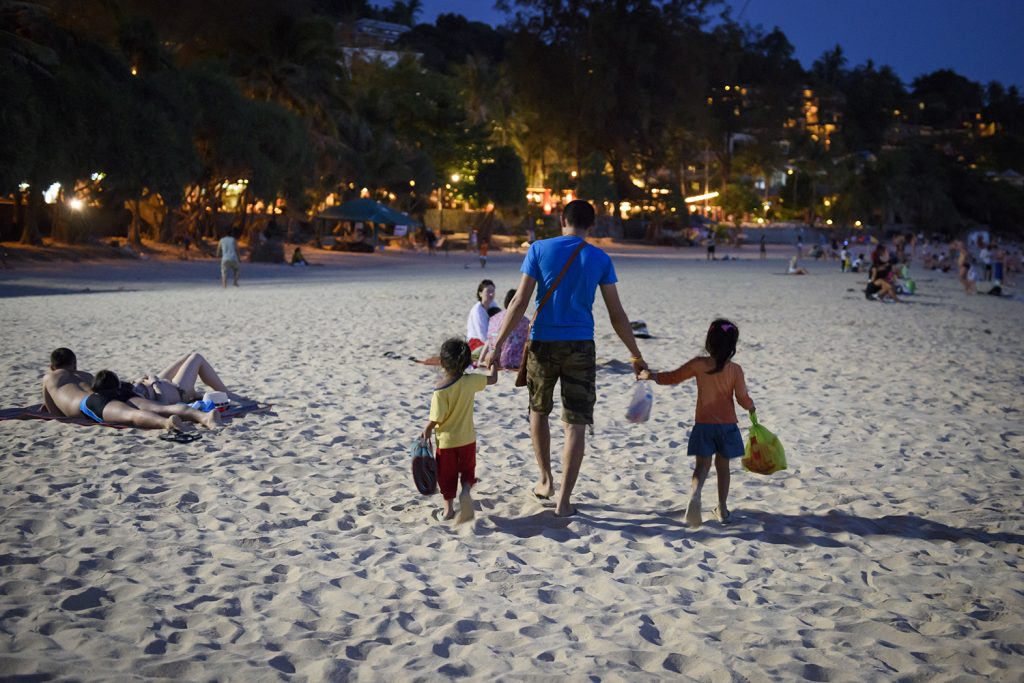 Två barn och en vuxen promenerar på en strand i Thailand.